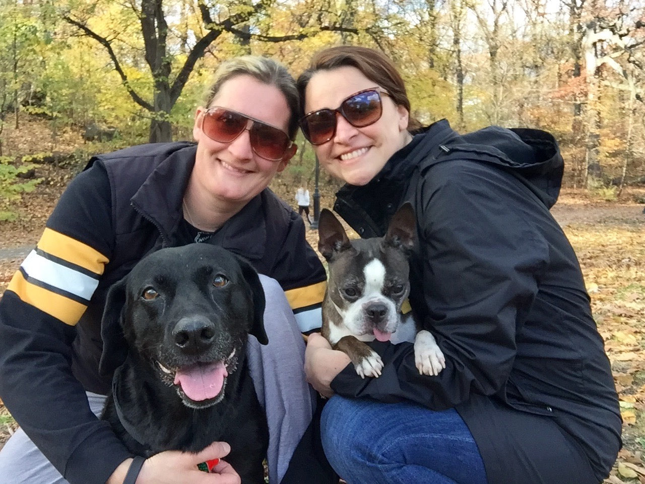 Co-founders of Wuffy, Ashley and Jaclyn pose with two dogs, Bailey and Gaia, outdoors in a park with autumn trees. 
