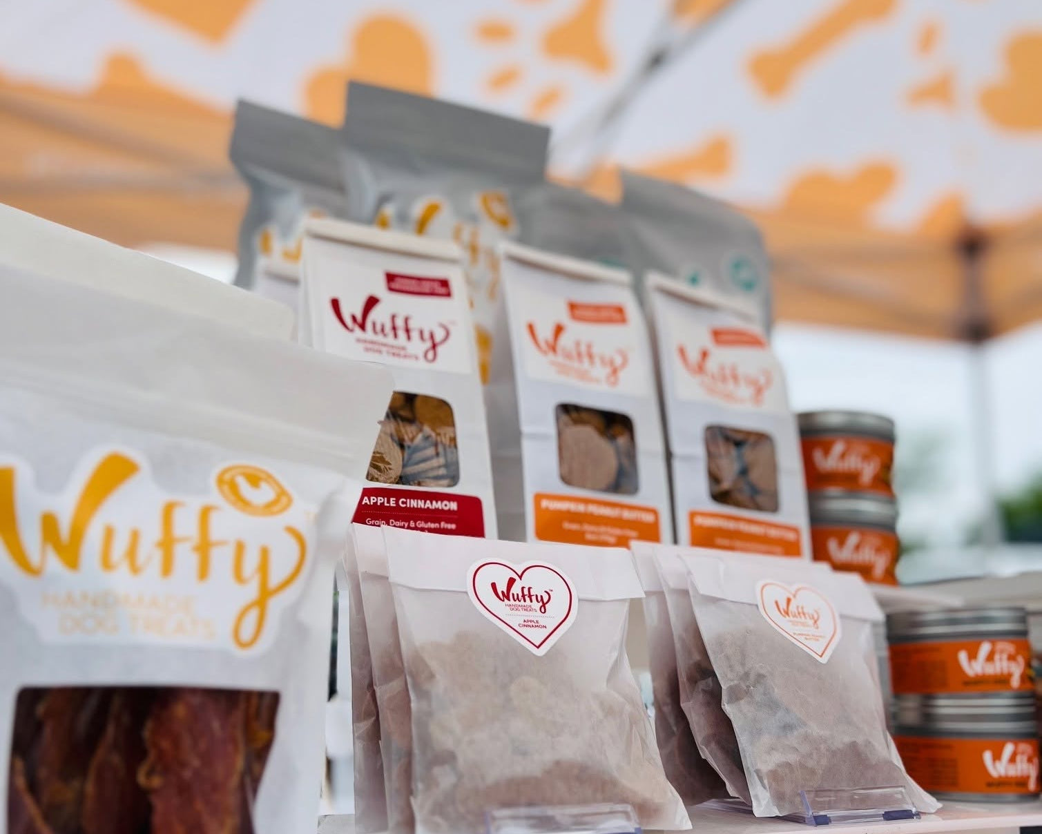 A display of Wuffy brand dog treats on a white table under an orange and white patterned canopy. In the foreground is a bag of Chicken Jerky, with several pouches of Apple Cinnamon and other flavors arranged behind it on a white stand.